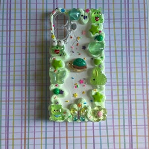 Peut inclure: Une coque de t&eacute;l&eacute;phone rectangulaire d&eacute;cor&eacute;e de bordures en cr&egrave;me fouett&eacute;e vert clair. La surface est orn&eacute;e de vermicelles color&eacute;s et de diverses breloques en r&eacute;sine, notamment une tortue, une grenouille, un lapin, des papillons et des &eacute;toiles.