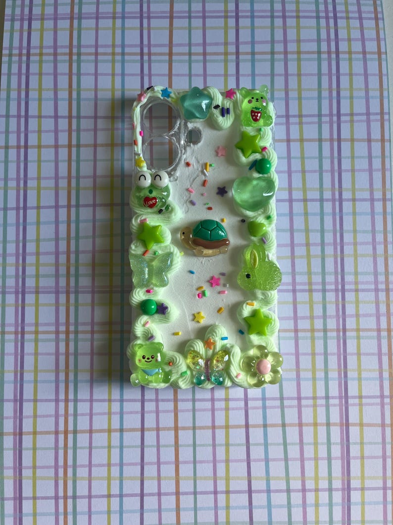 Peut inclure: Une coque de t&eacute;l&eacute;phone rectangulaire d&eacute;cor&eacute;e de bordures en cr&egrave;me fouett&eacute;e vert clair. La surface est orn&eacute;e de vermicelles color&eacute;s et de diverses breloques en r&eacute;sine, notamment une tortue, une grenouille, un lapin, des papillons et des &eacute;toiles.