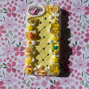 Peut inclure: Une coque de t&eacute;l&eacute;phone jaune d&eacute;cor&eacute;e d'une texture de cr&egrave;me fouett&eacute;e blanche, de strass incrust&eacute;s en losange et de divers charms jaunes translucides, notamment des ours, des &eacute;toiles, des c&oelig;urs, des papillons et un n&oelig;ud sur le bord sup&eacute;rieur.