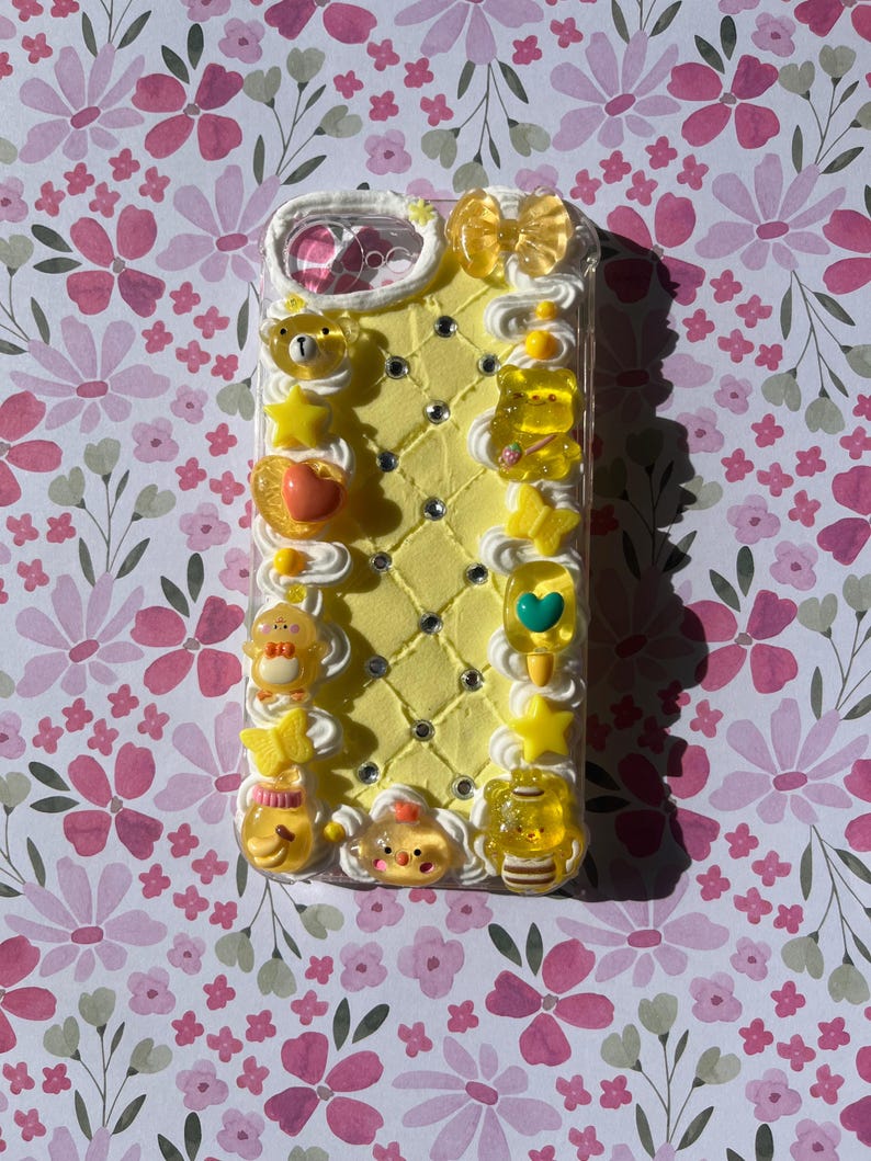 Peut inclure: Une coque de t&eacute;l&eacute;phone jaune d&eacute;cor&eacute;e d'une texture de cr&egrave;me fouett&eacute;e blanche, de strass incrust&eacute;s en losange et de divers charms jaunes translucides, notamment des ours, des &eacute;toiles, des c&oelig;urs, des papillons et un n&oelig;ud sur le bord sup&eacute;rieur.