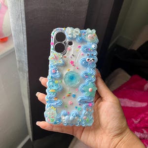 Peut inclure: Une coque de t&eacute;l&eacute;phone blanche d&eacute;cor&eacute;e de bordures bleues fa&ccedil;on gla&ccedil;age, de breloques color&eacute;es en forme d'&eacute;toiles et de vermicelles, ainsi que de petites figurines. Une fleur bleue est centr&eacute;e au dos et le mot love est visible.