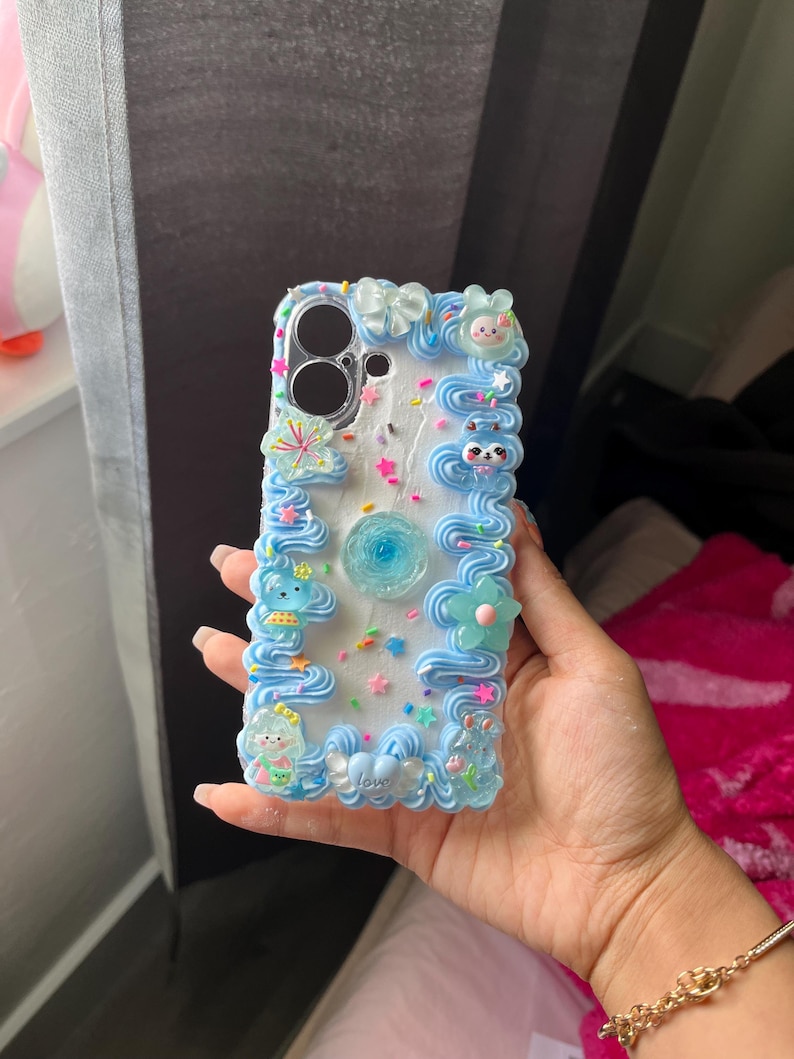 Peut inclure: Une coque de t&eacute;l&eacute;phone blanche d&eacute;cor&eacute;e de bordures bleues fa&ccedil;on gla&ccedil;age, de breloques color&eacute;es en forme d'&eacute;toiles et de vermicelles, ainsi que de petites figurines. Une fleur bleue est centr&eacute;e au dos et le mot love est visible.