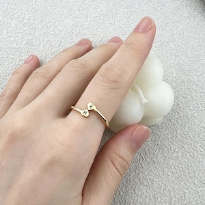 Peut inclure: Une bague ouverte dorée dotée de deux petites extrémités en forme de cœur, chacune gravée d'une lettre. Un cœur porte la lettre K et l'autre cœur porte la lettre H. La bague est portée sur un doigt, mettant en valeur son design délicat.