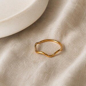 Puede incluir: Un anillo fino de color dorado con un diseño ondulado continuo y suave. La banda presenta un acabado metálico liso y pulido que refleja la luz, creando reflejos sutiles a lo largo de su forma orgánica y curvada.