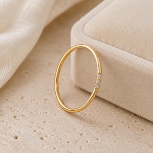 Puede incluir: Un anillo de banda fina de oro con un pequeño engaste rectangular incrustado con tres piedras diminutas, transparentes y de corte redondo. El metal tiene un acabado pulido y reflectante, con las piedras dispuestas en una línea vertical.