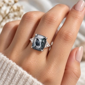 May include: A silver ring featuring a large rectangular gray stone with a detailed black raven illustration inside. The center stone is flanked by two small triangular clear stones on each side, all set in a polished silver band with prongs.