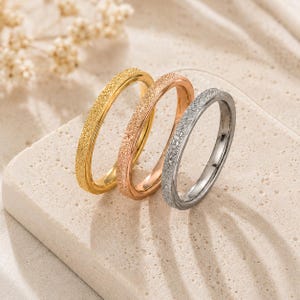 Puede incluir: Tres anillos de metal delgados dispuestos en fila, con una superficie texturizada y brillante. Los anillos tienen tres tonos metálicos distintos: oro amarillo brillante, oro rosa cálido y plata fría, cada uno con un ancho de banda uniforme.