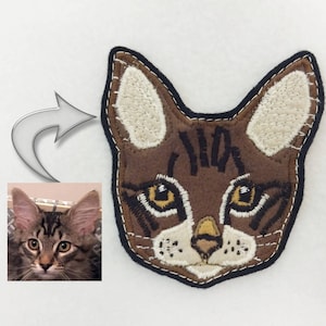 May include: A brown and white embroidered cat head patch with black stitching. The cat has yellow eyes and a white nose.