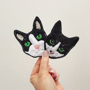 May include: Two black and white cat face patches with green eyes. The patches are made of felt and are embroidered with details.