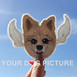 May include: A brown and white embroidered dog patch with angel wings. The patch is held in a hand against a blue sky background. The text "YOUR DOG PICTURE" is visible below the patch.