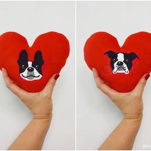 May include: Two red felt heart-shaped pillows with a black and white dog face embroidered on each. The dog has a white muzzle and black eyes.