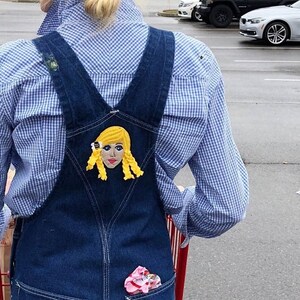 Puede incluir: Una persona lleva un mono vaquero azul con una camisa a cuadros blancos y azules. El mono tiene un parche en la espalda con una imagen bordada de una mujer rubia con una flor amarilla en el pelo.