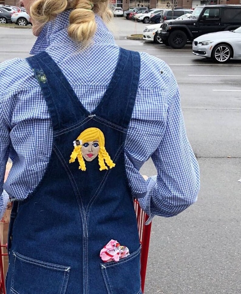 Puede incluir: Una persona lleva un mono vaquero azul con una camisa a cuadros blancos y azules. El mono tiene un parche en la espalda con una imagen bordada de una mujer rubia con una flor amarilla en el pelo.