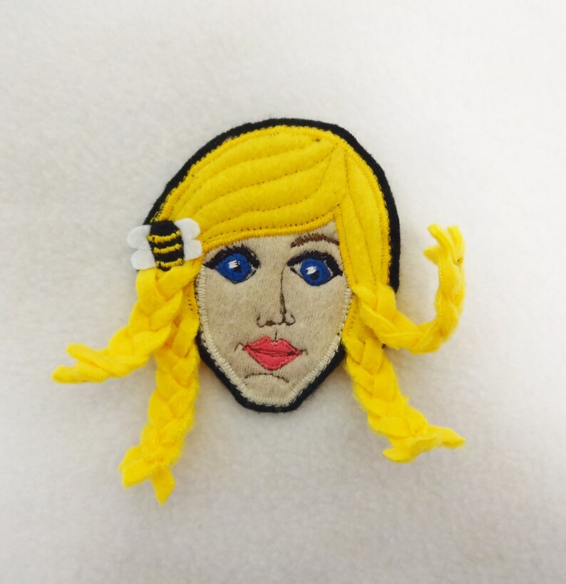 Puede incluir: Una aplicaci&oacute;n de fieltro de una ni&ntilde;a con trenzas rubias, ojos azules y una pinza para el pelo en forma de abeja. La aplicaci&oacute;n est&aacute; hecha de fieltro amarillo, negro, blanco y marr&oacute;n.