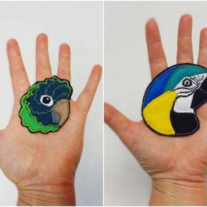 May include: Two colorful parrot head patches. One patch features a green and blue parrot head with a tan beak. The other patch shows a blue, yellow, and black parrot head with a white face and a black beak. Both patches are made of felt.