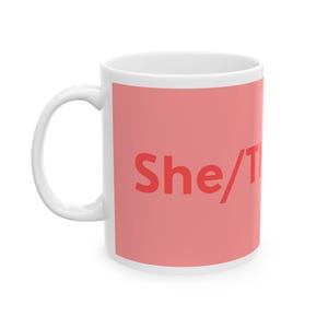 May include: A white ceramic mug featuring a large, rectangular coral-pink graphic panel on the side. The text She/They is printed in a bold, red sans-serif font across the center of the pink area. The mug has a curved handle on the left side.