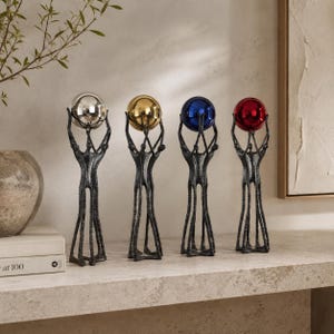 May include: Four tall, dark gray textured metal sculptures of stylized figures standing together, each holding a smooth, reflective sphere above their heads. The spheres are colored silver, gold, blue, and red, creating a vibrant contrast.