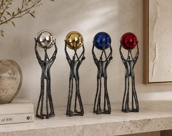 May include: Four tall, dark gray textured metal sculptures of stylized figures standing together, each holding a smooth, reflective sphere above their heads. The spheres are colored silver, gold, blue, and red, creating a vibrant contrast.