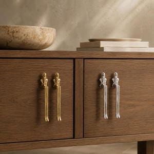 May include: Four metal cabinet pulls shaped like stylized figures sitting with long legs dangling down. Two pulls are finished in a polished gold tone, and two pulls are finished in a polished silver tone, all mounted on a dark wood cabinet.