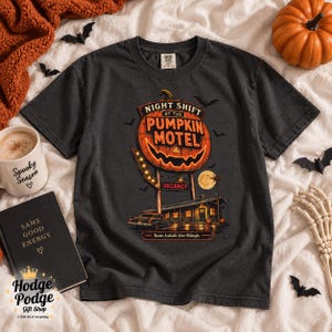 May include: A charcoal gray t-shirt featuring a graphic of a glowing jack-o'-lantern sign that reads Night Shift at the Pumpkin Motel, with a vintage car, a full moon, and the text Vacancy and Rooms Available After Midnight printed on the front.