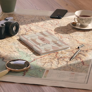Può includere: Un taccuino rettangolare con un motivo geometrico beige e marrone sulla copertina, che riporta il testo "Love... in every page". Il taccuino è posizionato su una mappa vintage insieme a una fotocamera nera, uno smartphone, una tazza da tè e una penna.