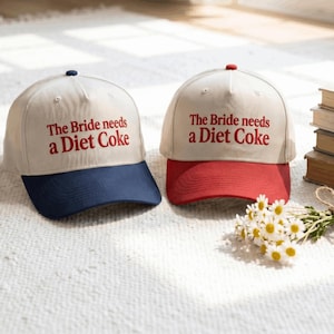 Może przedstawiać: Dwie czapki z daszkiem z kremowymi koronami i wyhaftowanym czerwonym napisem The Bride needs a Diet Coke. Jedna czapka ma granatowy daszek, a druga czerwony. Obie czapki mają pasujący kolorystycznie guzik na czubku głowy.