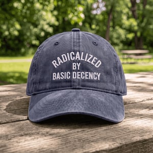 Könnte beinhalten: Eine marineblaue Baseballkappe mit verwaschenem, abgenutztem Finish. Auf der Vorderseite befindet sich eine weiße Stickerei mit dem Text RADICALIZED BY BASIC DECENCY, wobei das erste Wort in einem Bogen über den anderen Wörtern steht.