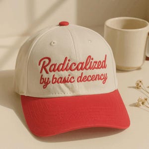 May include: A cream-colored baseball cap with a red brim and a red button on top. The front features the embroidered red text "Radicalized by basic decency" in a cursive script font. The cap has a structured crown with ventilation eyelets.