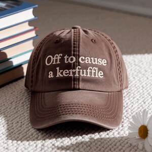 May include: A brown baseball cap with a curved brim and embroidered light-coloured text on the front that reads "Off to cause a kerfuffle". The cap features a six-panel construction with a button on top and stitched ventilation eyelets.