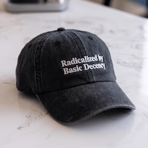 Puede incluir: Una gorra de béisbol gris carbón con un acabado desgastado y lavado. La parte frontal presenta un texto bordado en blanco que dice Radicalized by Basic Decency. La gorra tiene una corona estructurada, una visera curva y un botón superior.