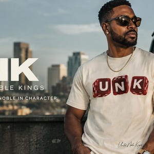 Puede incluir: Una camiseta de manga corta color crema con un gráfico de tres dados rojos que muestran las letras U, N y K. El texto UNK, UNITED NOBLE KINGS y YOUTHFUL IN SPIRIT. NOBLE IN CHARACTER está impreso en el lado izquierdo de la imagen.