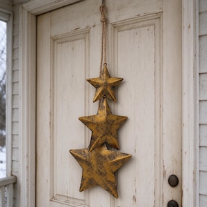 May include: A vertical wall hanging featuring three five-pointed stars in descending sizes, crafted from a metallic gold material with a dark, distressed, weathered finish, suspended from a single thick, natural-colored jute rope loop.