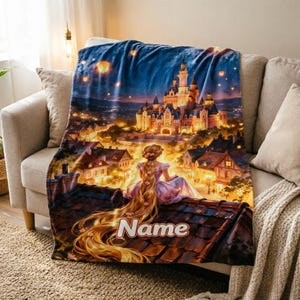Pode incluir: Uma manta de lã macia com uma cena impressa de uma personagem de cabelos longos e loiros sentada num telhado, com vista para um castelo e uma cidade iluminados à noite, com lanternas flutuantes e a palavra Name impressa nas telhas.