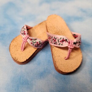 May include: A pair of miniature flip-flops with light brown soles and pink straps. The straps are adorned with a floral pattern in shades of pink, green, and white. The flip-flops are designed for a doll or small figurine.