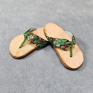 May include: A pair of flip-flops with a cork footbed and a camouflage pattern strap. The straps are green and brown, and the thong is green. The flip-flops are on a gray surface.