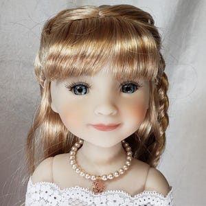 May include: A doll with long, wavy, blonde hair, blue eyes, and a pink and gold necklace. The doll is wearing a white lace dress.