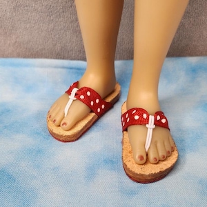 May include: Red and white polka dot flip-flops with a white thong strap. The sandals have a cork-like sole and are worn by a doll. The red strap is a ribbon-like material.