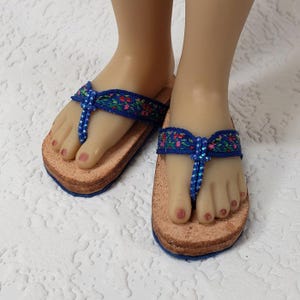 May include: A pair of miniature doll sandals. The sandals have a cork footbed with a blue trim. The straps are blue with a floral pattern and a beaded thong. The doll's feet are visible.