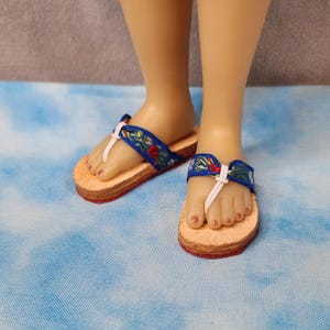 May include: A pair of miniature doll sandals. The sandals have a cork sole with a red edge and a blue fabric strap embroidered with flowers. A white thong strap secures the sandal to the foot.