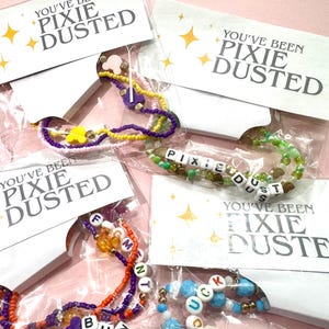 May include: Four beaded bracelets in clear plastic packaging, each attached to a white card printed with the text YOU'VE BEEN PIXIE DUSTED and yellow star icons. The bracelets feature colorful seed beads, letter cubes, and small mouse-shaped charms.