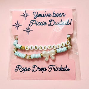 May include: Two beaded bracelets on a pink display card labeled You've been Pixie Dusted! and Rope Drop Trinkets. One bracelet features white beads with gold letters spelling CINDERELLA, while the other includes light blue disc beads and a white bow.