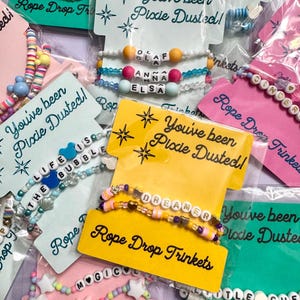 May include: A collection of beaded bracelets packaged on individual display cards. Each card features the text 'You've been Pixie Dusted!' and 'Rope Drop Trinkets'. The bracelets include colourful plastic beads, letter beads spelling names or phrases, and small character-shaped charms.