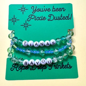 May include: Three beaded bracelets on a green display card with the text You've been Pixie Dusted! and Rope Drop Trinkets. The bracelets feature clear green crackle beads, blue faceted beads, and white letter beads spelling TIANAS BAYOU.