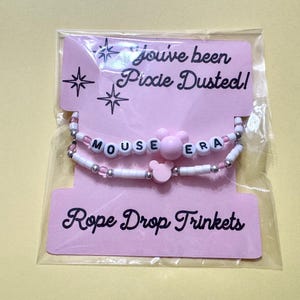 May include: Two beaded bracelets on a pink display card labeled You've been Pixie Dusted! and Rope Drop Trinkets. The top bracelet features white letter beads spelling MOUSE ERA with a pink mouse-shaped bead. The bottom has white and silver beads.