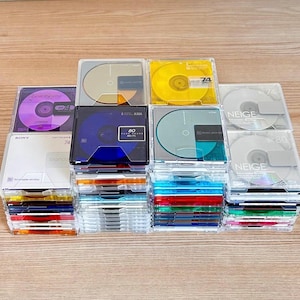 May include: A collection of square MiniDisc cases stacked in multiple columns. The cases are transparent plastic, revealing colorful discs in shades of purple, gold, yellow, blue, green, and silver. Some labels feature text including Sony, Axia, and Neige.