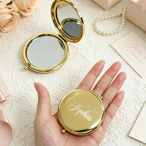 May include: A round, gold-toned metal compact mirror with a polished finish. One mirror is open, revealing two reflective surfaces, while the other is closed and features the name Sophia engraved in a white script font on the front cover.