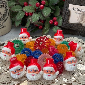 May include: A collection of vintage Christmas light covers shaped like Santa Claus faces with red hats and white beards. The lights are surrounded by colorful plastic reflectors and sit on a decorative silver tray.