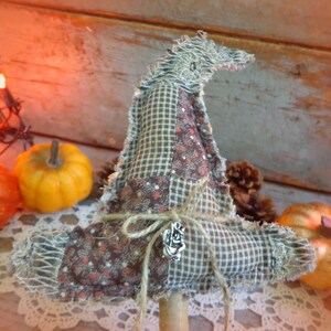 May include: A fabric witch hat featuring a patchwork design of brown and tan checkered fabric and brown floral print fabric. The hat has frayed edges, a pointed top, and is tied with a piece of twine featuring a small silver-toned metal charm.