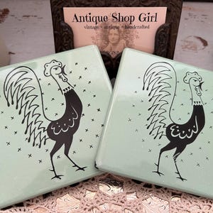 May include: Two square, light green ceramic coasters with a black rooster design. The rooster is stylized with a long neck and detailed tail feathers. Small black crosses are scattered across the surface. The coasters are displayed on a decorative surface.