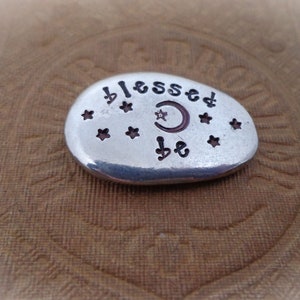May include: A silver-colored, oval-shaped stone with the words "blessed be" and star and crescent moon designs. The stone is set against a textured, brown background.